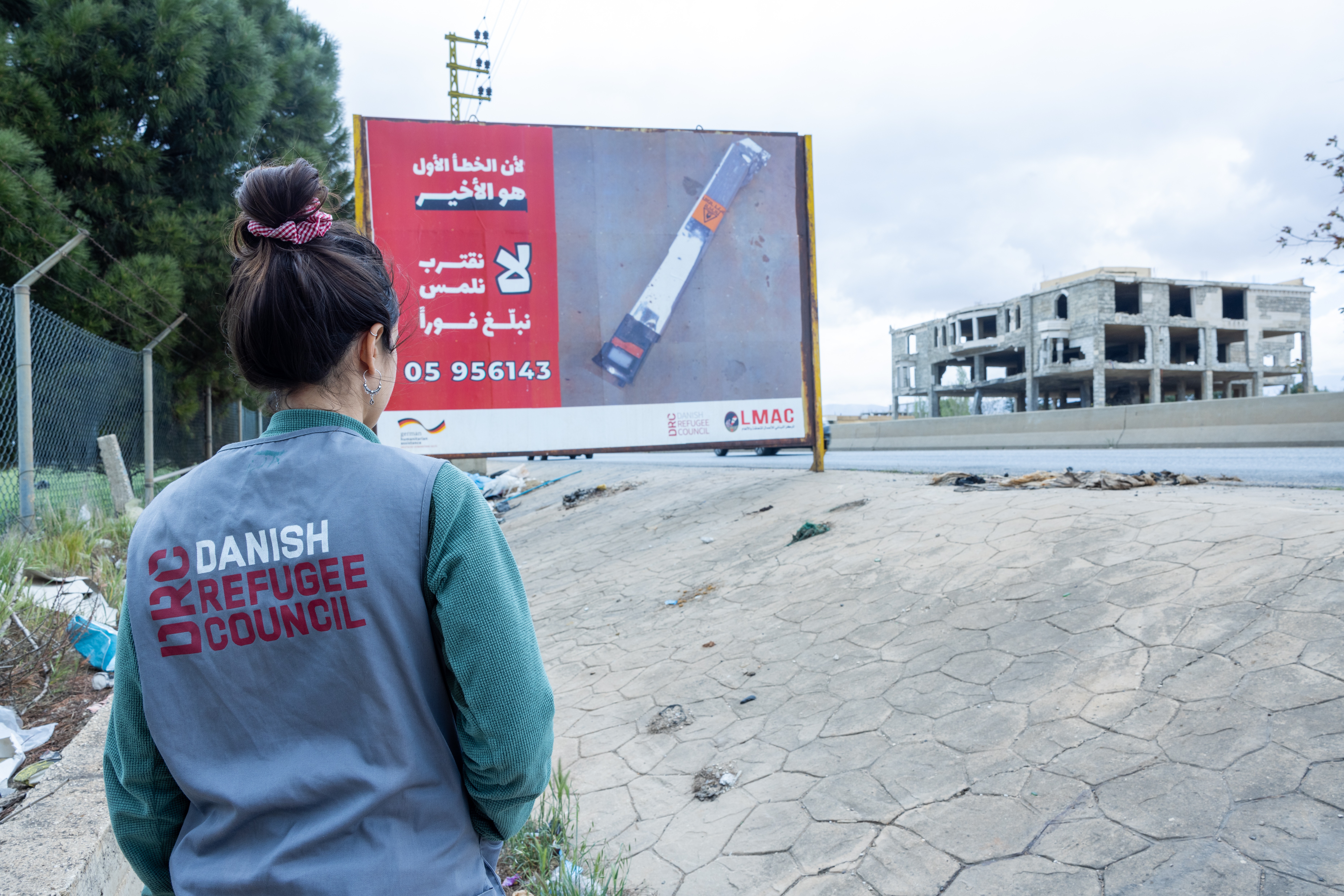 A memebr of DRC staff in Lebanon stands in front of a poster that educate people on the risk of explosive ordnance